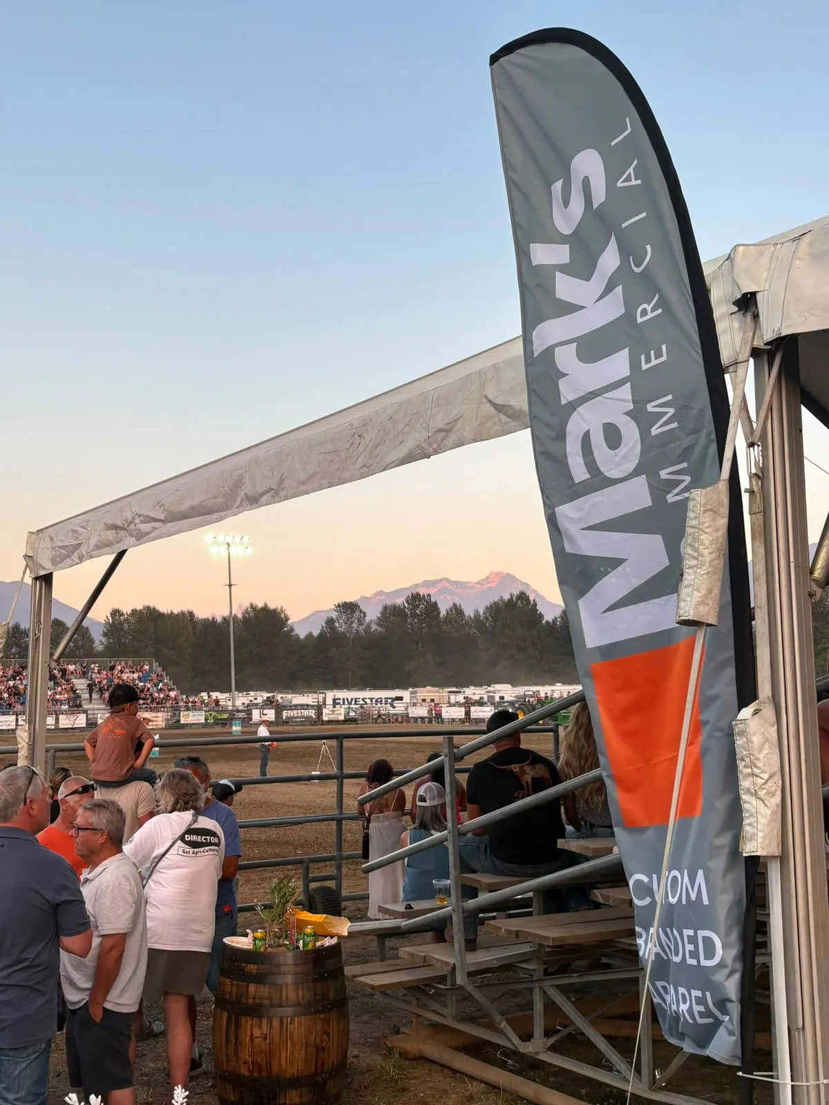 Mark's Commercial custom branded apparel banner at the 2026 Chilliwack Fair, grandstand and Fraser Valley mountains behind it at sunset